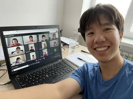 A volunteer smiling beside a laptop that shows an online lesson with about a dozen students on video.