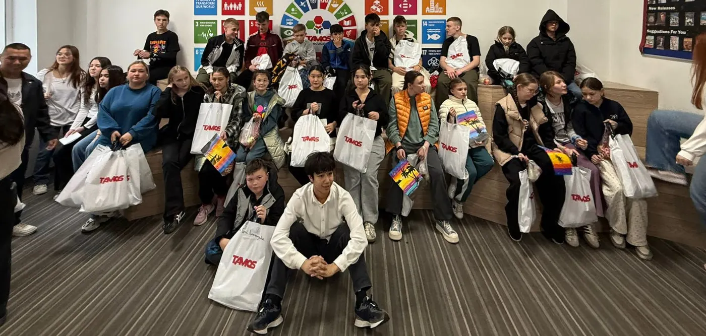 About thirty children holding gift bags and paintings, seated on tiered benches in a classroom at Tamos Education.